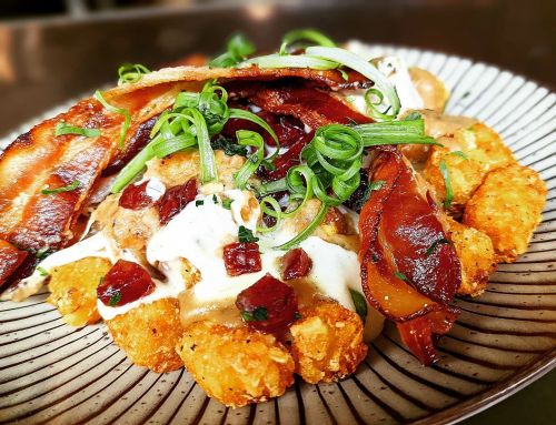A plate of loaded tater tots topped with bacon, green onions, and a creamy sauce, garnished with herbs.