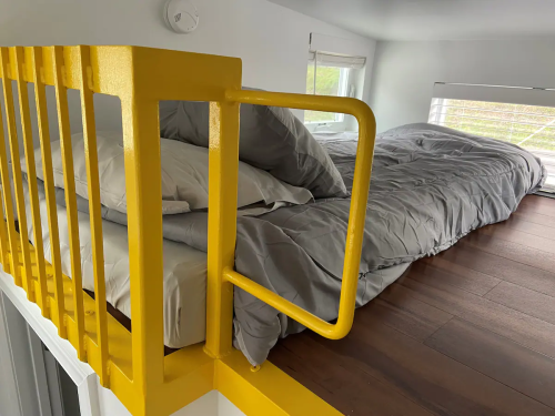 A cozy loft bed with gray bedding, featuring a bright yellow railing and large windows for natural light.