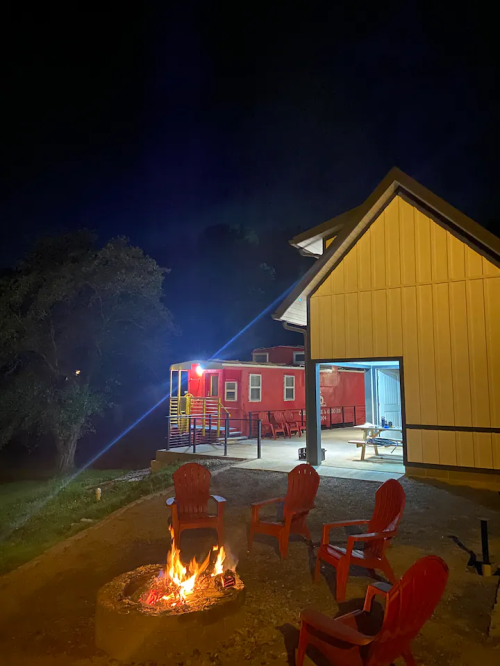 A cozy outdoor scene at night featuring a fire pit, red chairs, and a brightly lit building in the background.