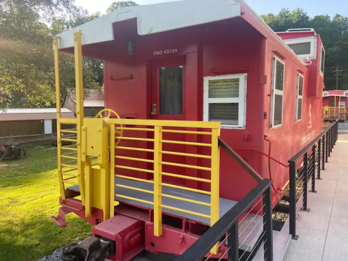 A bright red caboose with yellow accents, featuring a railing and steps, set in a grassy area.