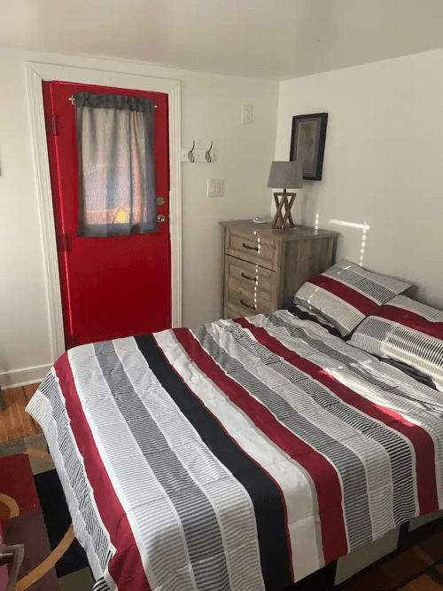 A cozy bedroom featuring a striped bedspread, a wooden dresser, and a bright red door with a curtain.