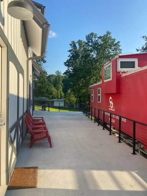 A red train caboose beside a modern building, with a patio area and trees in the background under a clear blue sky.