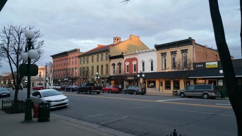 Historic buildings line a street at dusk, with cars parked and streetlights illuminating the scene.