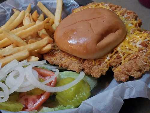 A plate with a large fried chicken sandwich topped with cheese, served with fries, lettuce, tomato, pickles, and onions.