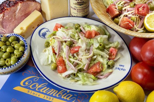 A fresh salad with lettuce, tomatoes, olives, and ham, surrounded by cheese, olives, and lemons on a colorful table.
