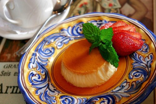A creamy flan topped with caramel, garnished with mint leaves and strawberries, served on a decorative blue plate.