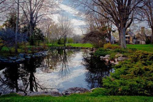 A serene landscape featuring a pond reflecting trees and a house, surrounded by lush greenery and rocks.
