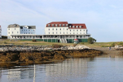 A large white hotel with red roofs overlooks a calm water body, surrounded by grassy land and rocky shoreline.