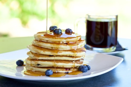 A stack of pancakes drizzled with syrup and topped with blueberries, alongside a cup of coffee.