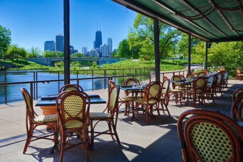 Outdoor dining area with empty tables overlooking a river and city skyline on a sunny day.