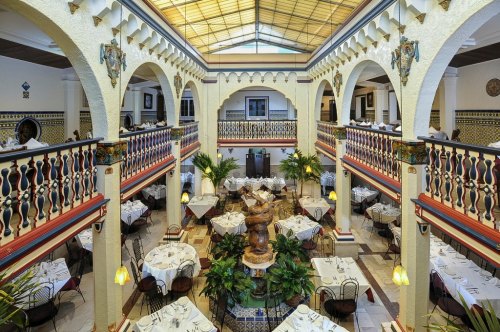 A spacious restaurant interior with arched balconies, elegant tables, and a central fountain surrounded by greenery.