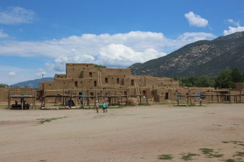 A traditional adobe pueblo structure with a mountainous backdrop and a clear blue sky. Two people walk in the foreground.