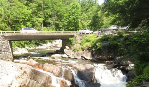 A bridge spans a rocky river surrounded by lush green trees, with cars parked nearby.