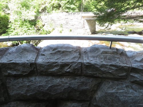 A stone wall with a metal railing overlooks a river and a bridge surrounded by lush greenery.