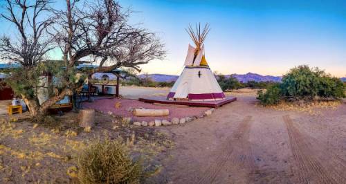 A scenic view of a teepee surrounded by desert landscape, with mountains in the background and a clear blue sky.