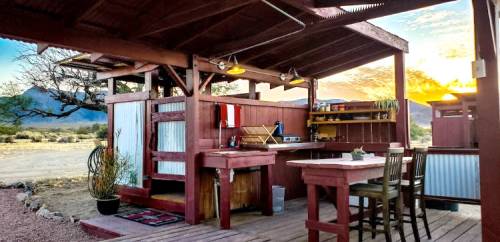 Cozy outdoor kitchen with wooden furniture, sink, and shelves, set against a desert landscape at sunset.