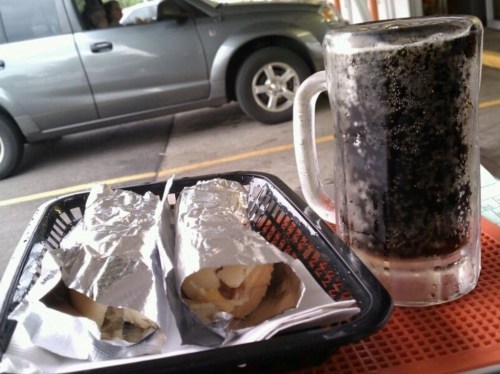 A tray with two foil-wrapped burritos and a frosty mug of dark beer, with a car visible in the background.