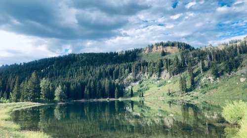 A serene lake surrounded by lush green hills and trees, reflecting the cloudy sky above.
