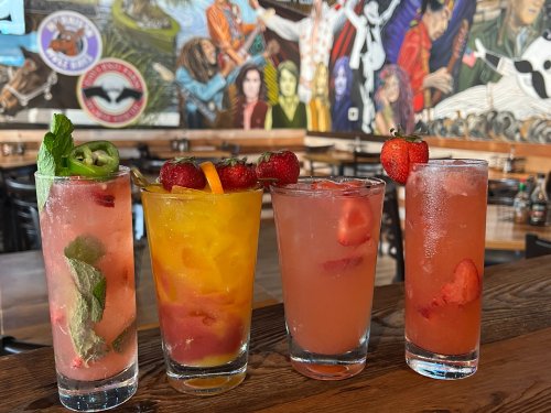 Four colorful cocktails with fresh fruit garnishes on a wooden table, set against a vibrant mural backdrop.