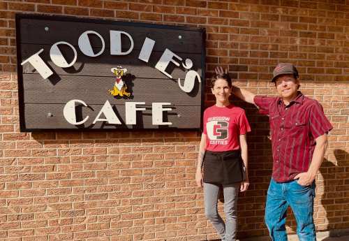 A man and woman stand beside the Toodie's Cafe sign, smiling in front of a brick wall.