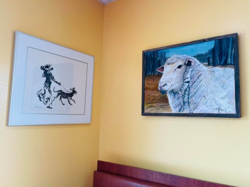 Two framed artworks on a yellow wall: a black-and-white drawing of dogs and a colorful painting of a sheep.