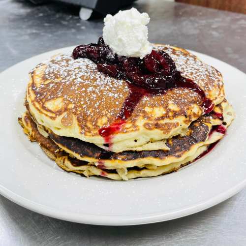 Stack of fluffy pancakes topped with whipped cream and berry sauce, dusted with powdered sugar on a white plate.
