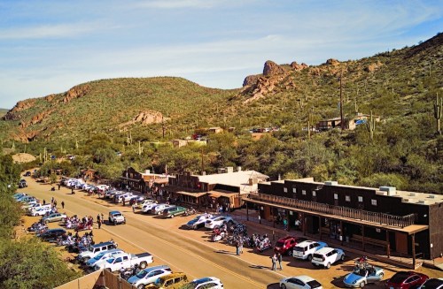 Aerial view of a small town with parked cars and motorcycles, surrounded by mountains and desert vegetation.