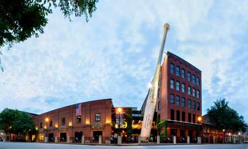 A large baseball bat leans against a brick building, with a cloudy sky above and lights illuminating the scene.