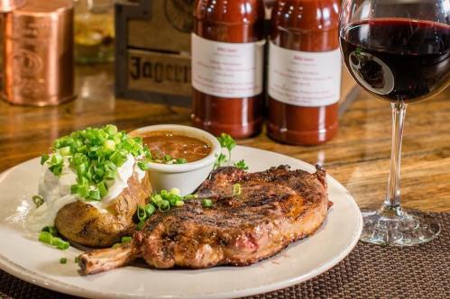 A plate featuring a grilled pork chop, baked potato with sour cream and green onions, barbecue sauce, and a glass of red wine.