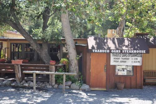 Exterior of Trabuco Oaks Steakhouse with wooden signage, surrounded by trees and gravel, inviting atmosphere.