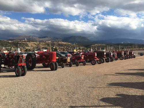 A row of vintage red tractors lined up on a gravel surface with mountains and cloudy skies in the background.