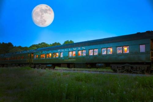 A vintage Pullman train car under a bright full moon, surrounded by trees and a clear blue sky.