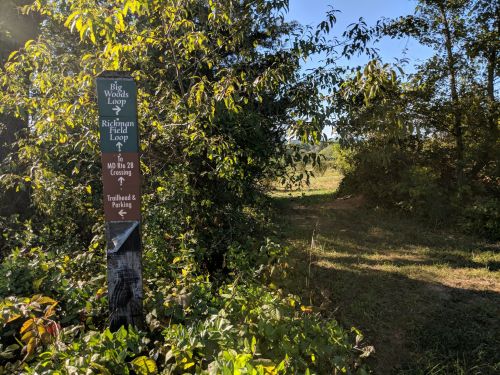 Trail sign indicating the Big Woods Loop and Richman Field Loop, surrounded by lush greenery and a clear path ahead.