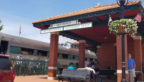 A train station with a sign for the Van Buren Visitors Center and Excursion Train, featuring people and hanging flower baskets.