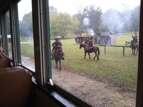 Two horse riders in cowboy attire on a grassy field, with smoke in the background and a building nearby.