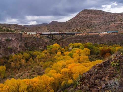 A train crosses a bridge over a canyon filled with vibrant autumn foliage and rugged mountains in the background.