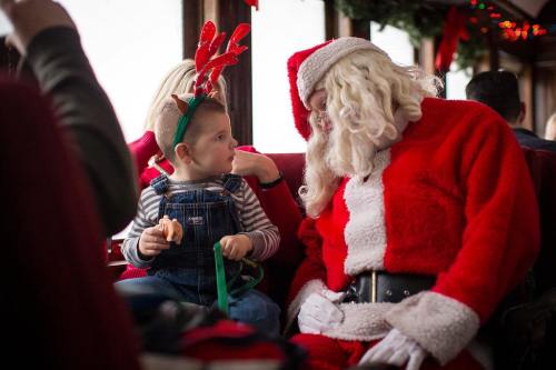 A child in reindeer antlers sits with Santa Claus on a festive train, sharing a joyful moment together.