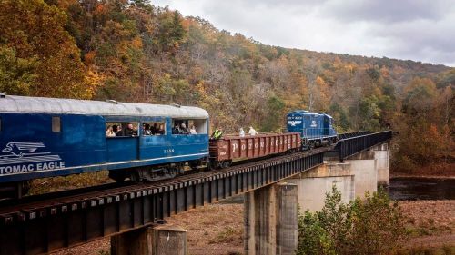 A blue train crosses a bridge over a river, surrounded by autumn foliage and rolling hills. Passengers can be seen inside.