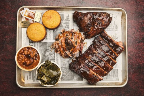 A metal tray with barbecue ribs, pulled pork, cornbread, baked beans, and collard greens on newspaper.
