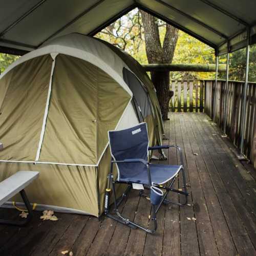 A camping tent set up on a wooden deck surrounded by trees, with a folding chair nearby.