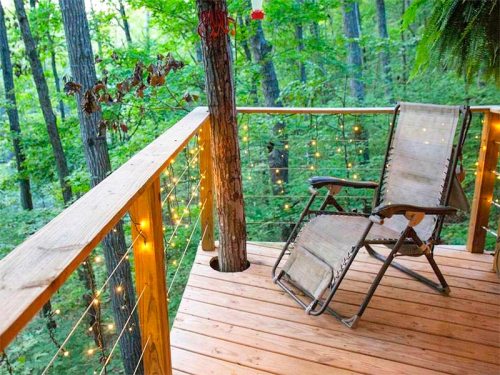 A wooden deck in a forest, featuring a lounge chair and string lights, surrounded by lush green trees.