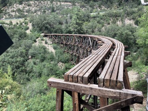 A wooden railway trestle bridge curves over a lush green landscape, surrounded by trees and hills.