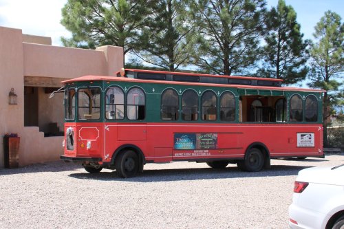 A red and green trolley bus parked outside a building, surrounded by trees and gravel.