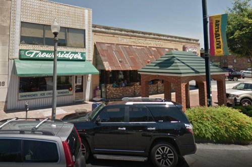 A street view of a restaurant with outdoor seating, surrounded by parked cars and a banner in the background.