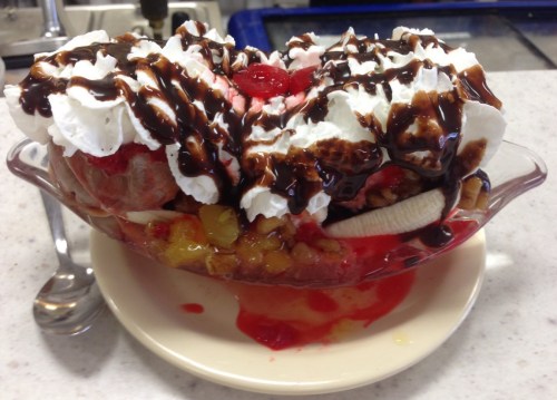 A colorful ice cream sundae topped with whipped cream, chocolate sauce, cherries, and fruit in a glass dish.