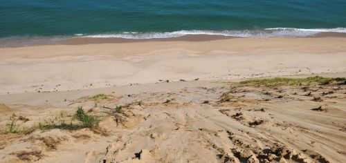 A sandy dune overlooking a calm blue ocean and a beach with gentle waves lapping at the shore.