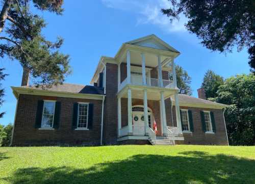 A historic two-story brick house with a front porch, surrounded by green grass and trees under a clear blue sky.