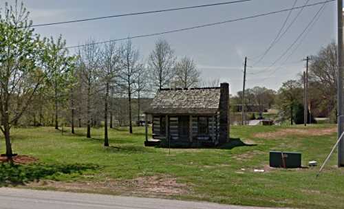 A small log cabin surrounded by trees and grass, with power lines in the background on a sunny day.