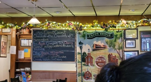 A cozy café interior with a chalkboard menu and festive decorations, featuring cakes and pies listed.