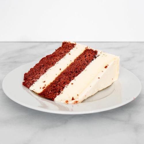 A slice of red velvet cake with cream cheese frosting on a light gray plate, set against a white background.